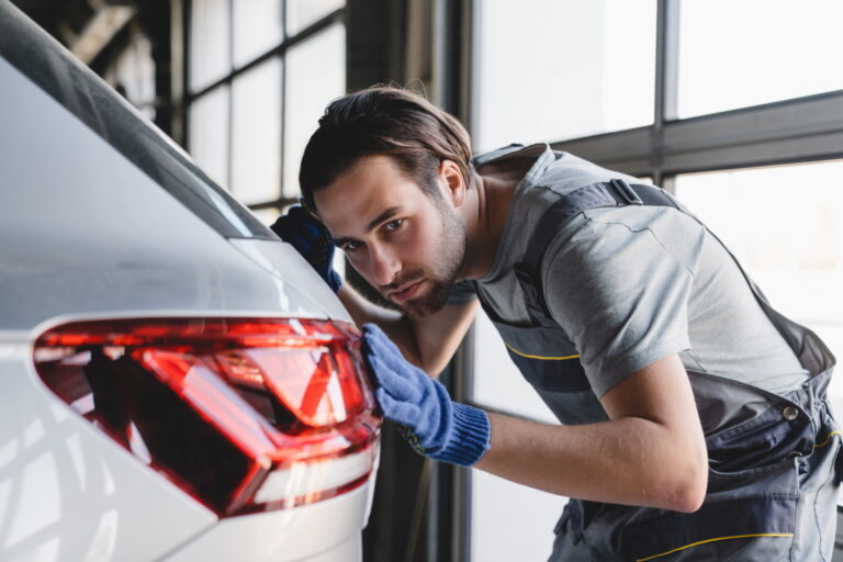 Caucasian male young technician mechanic in special robe uniform checking car paint repairing fixing after scratches at vehicle inspection service MOT.