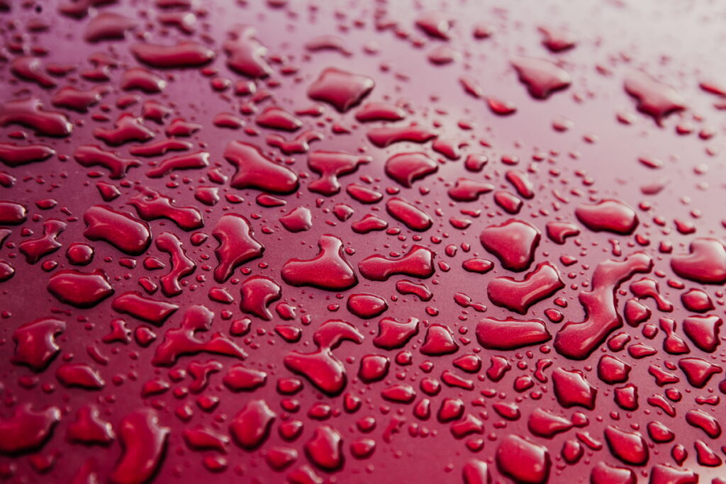 Water drops on clean red car. Abstract blur red background. Roof of car with wet surface. Realistic image. Rainstrorm. Rainy weather. Water droplets on metallic surface